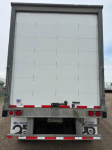 Rear view of a Wabash trailer with a white closed cargo door, red and white reflective tape, and bumper lights. VIN and stock not visible on door, but license plate area shows State plates and bumper equipment.