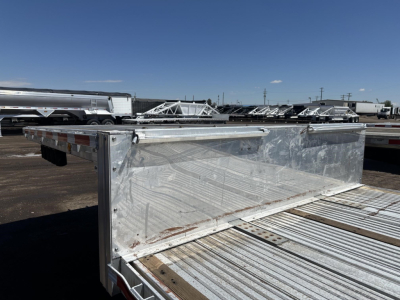 All-aluminum drop deck trailer viewed from side with front rack and ribbed deck, reflective metal surface, other trailers in background.