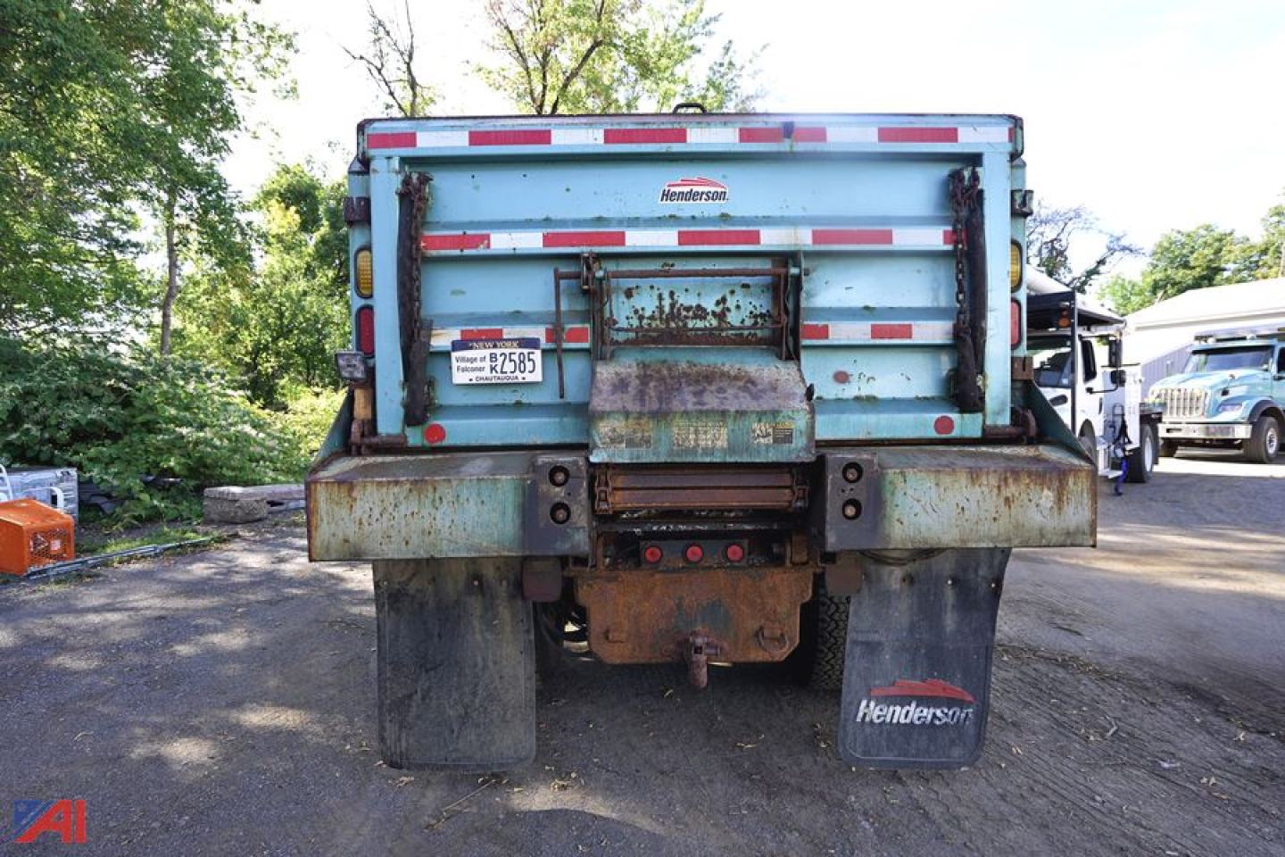 Dump truck rear showing rusted tailgate and hitch with Henderson mud flaps and spare tires in yard.