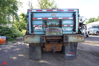 Dump truck rear showing rusted tailgate and hitch with Henderson mud flaps and spare tires in yard.