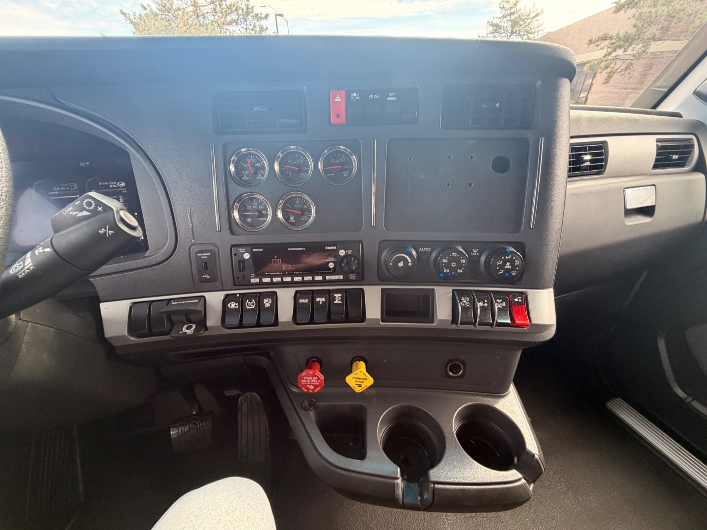 Dashboard and center console of a 2023 Kenworth T680 with steering column stalks, multiple round gauges, switches, and two cup holders in the lower dash area.