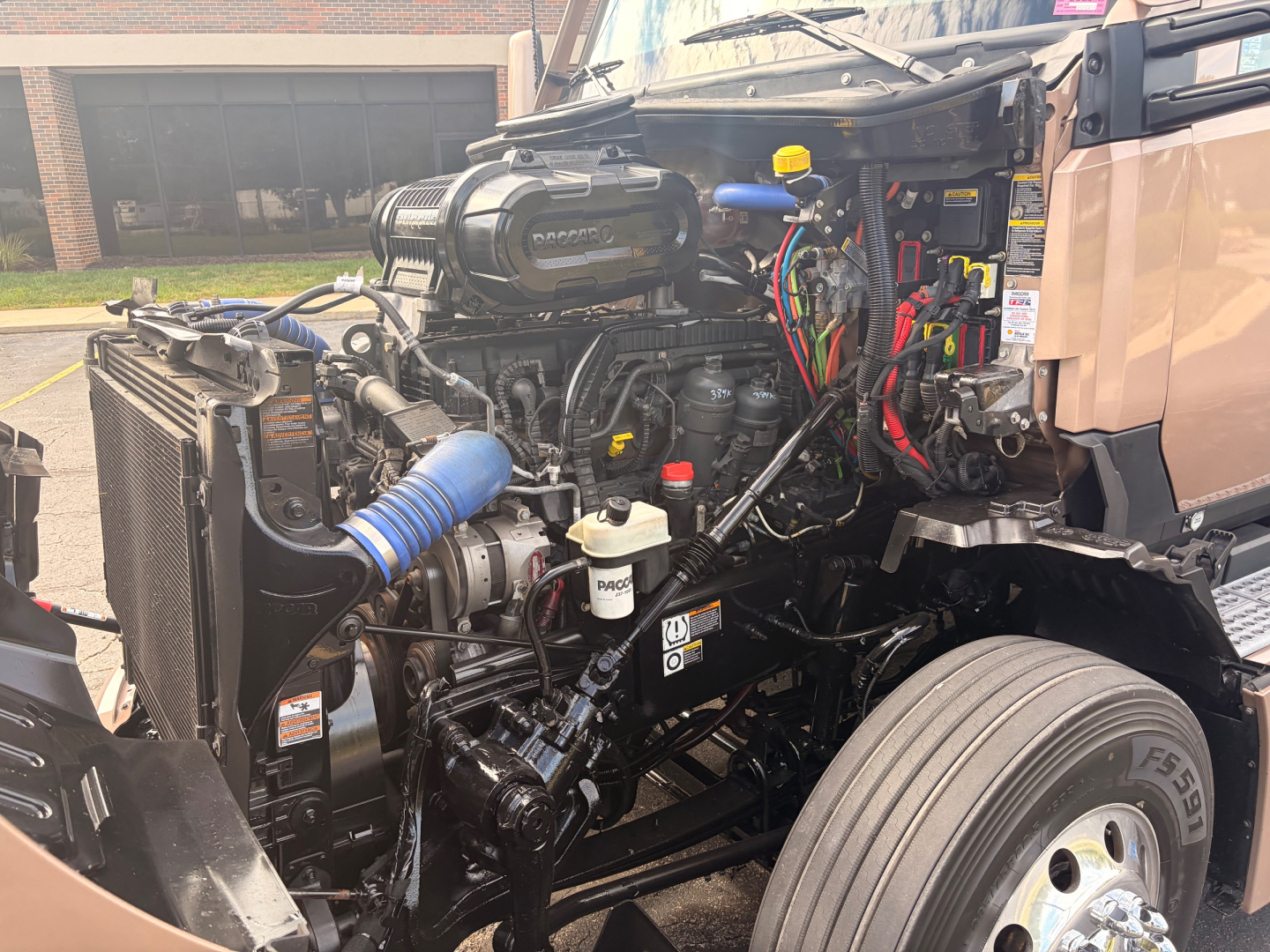 Kenworth T680 engine bay showing exposed diesel engine, hoses, wiring, coolant reservoir, and front wheel in view.