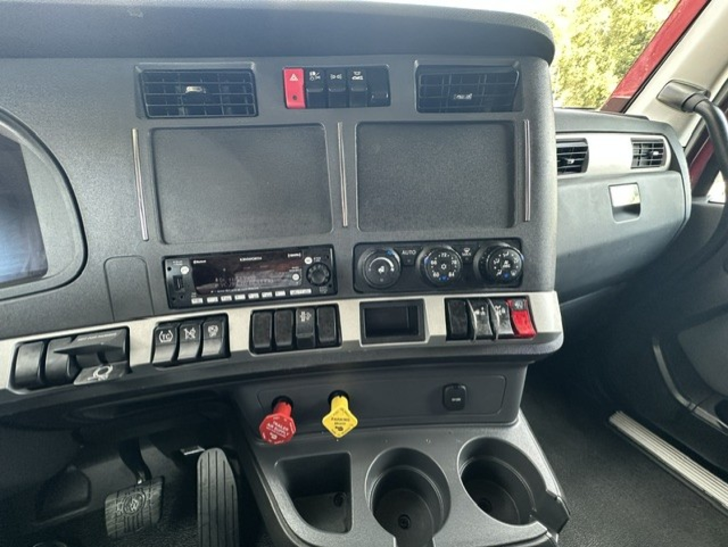 Kenworth T680 dash view showing two large blank screens, climate controls, radio, various switches, red hazard button, yellow and red handles, cup holders, and dashboard area.