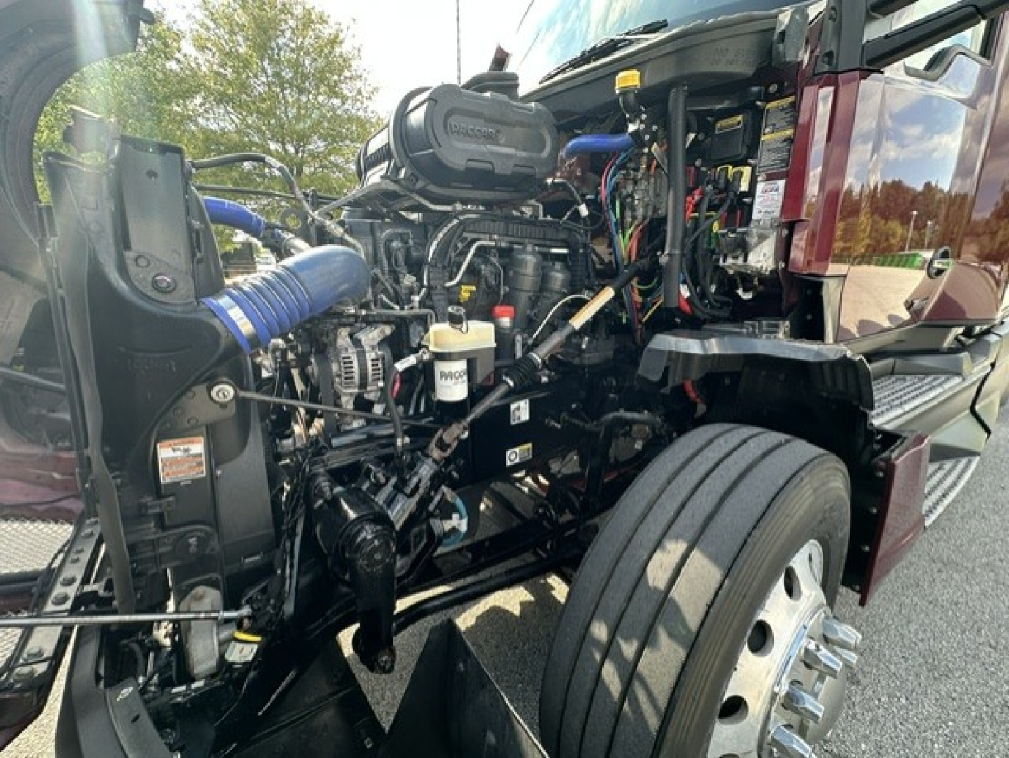 Kenworth T680 engine bay with exposed diesel engine, hoses, and components visible; blue intake hose, yellow cap, wiring, and part labels.