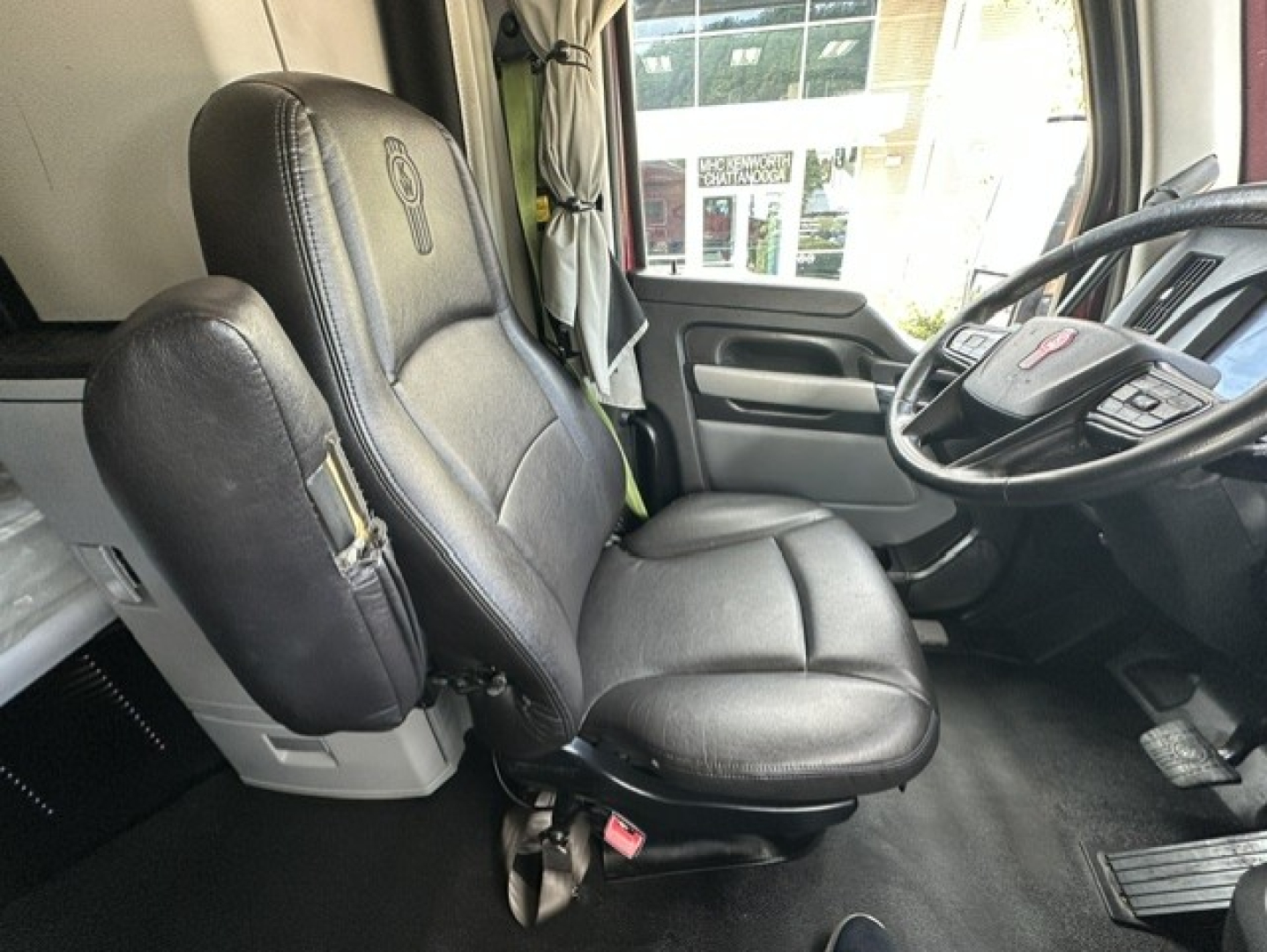 Kenworth T680 cab interior showing driver seat and steering wheel from the passenger side. Leather seat, dashboard, pedals visible.