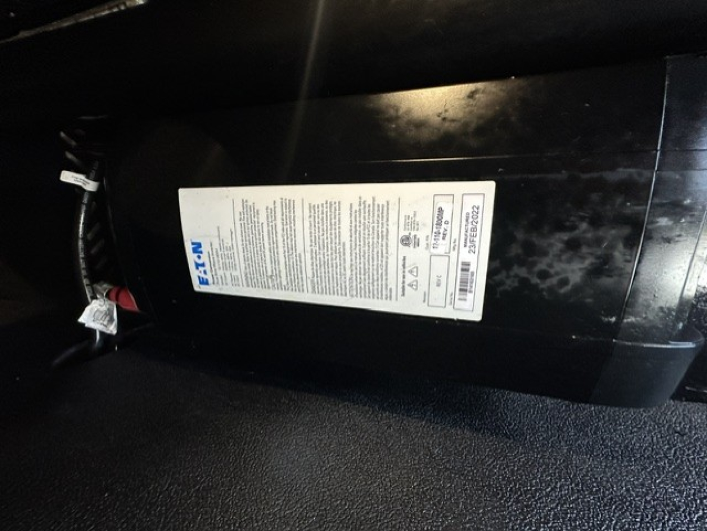 Kenworth T680 under-hood view showing a rectangular white label with vehicle specifications and a black cylindrical component, wiring harness visible.