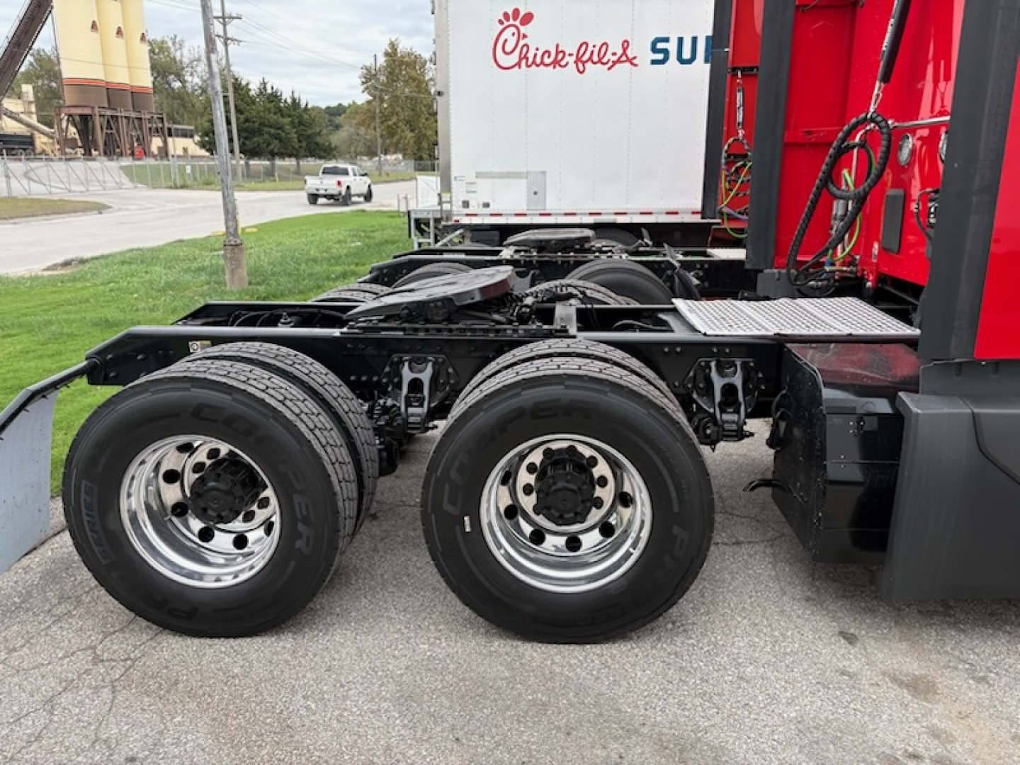 Kenworth T680 tractor chassis showing dual rear axles with black tires and chrome rims, behind the cab with red painted body and a white Chick-fil-A trailer in background