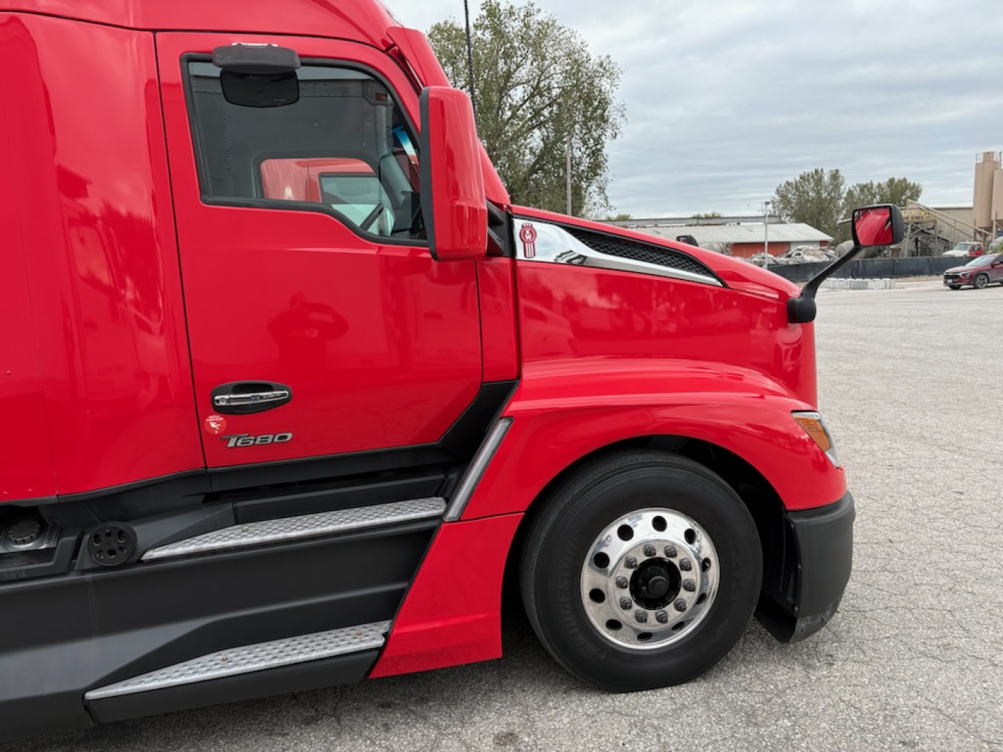 Truck cab side of a red 2023 Kenworth T680 with chrome wheel, black lower body trim, and side steps visible.