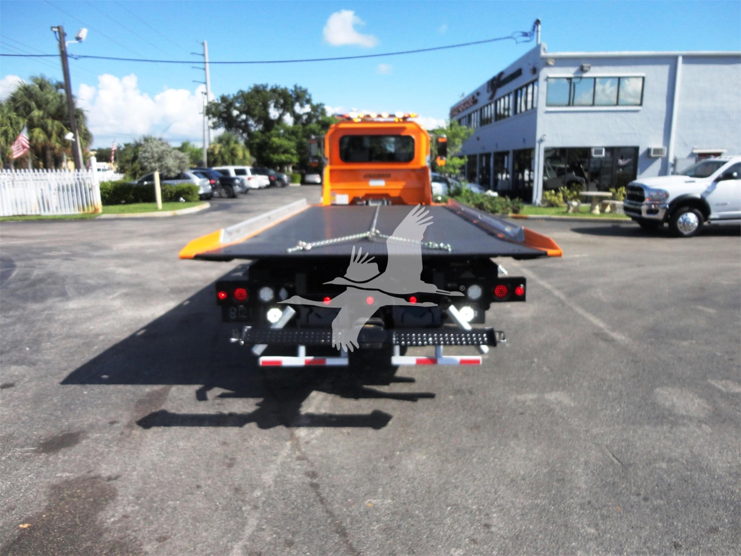Back of a 2022 Peterbilt 337 rollback truck with a flatbed; visible are orange cab, rear deck, taillights, and a building in the background.
