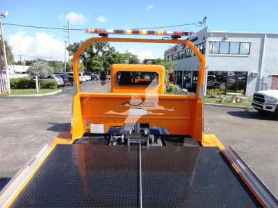 Peterbilt 337 rollback truck bed with bright orange chassis and reflective safety bar; rear view showing flatbed surface and hoist area, dealership lot in background.