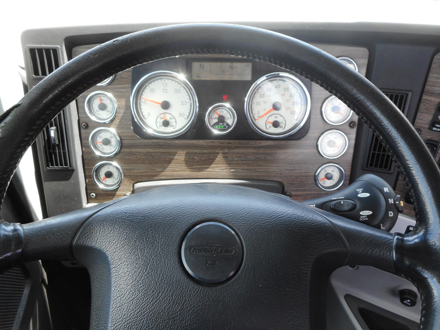 Dashboard view of a Freightliner 122SD truck cab showing gauges, steering wheel with Freightliner logo, woodgrain dash trim, and control stalks.
