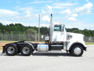 Side view of a white Freightliner 122SD day cab truck with chrome exhaust, fuel tanks, and triple rear axles.