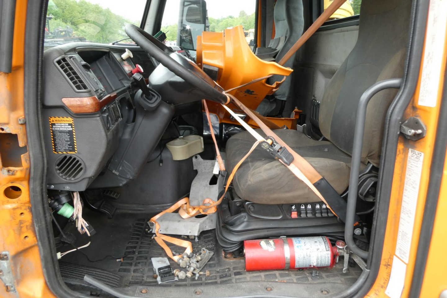 Truck cab interior showing steering wheel, dashboard, driver's seat with seatbelt, orange equipment mounted near seat, scattered tools, and a red fire extinguisher on the floor.