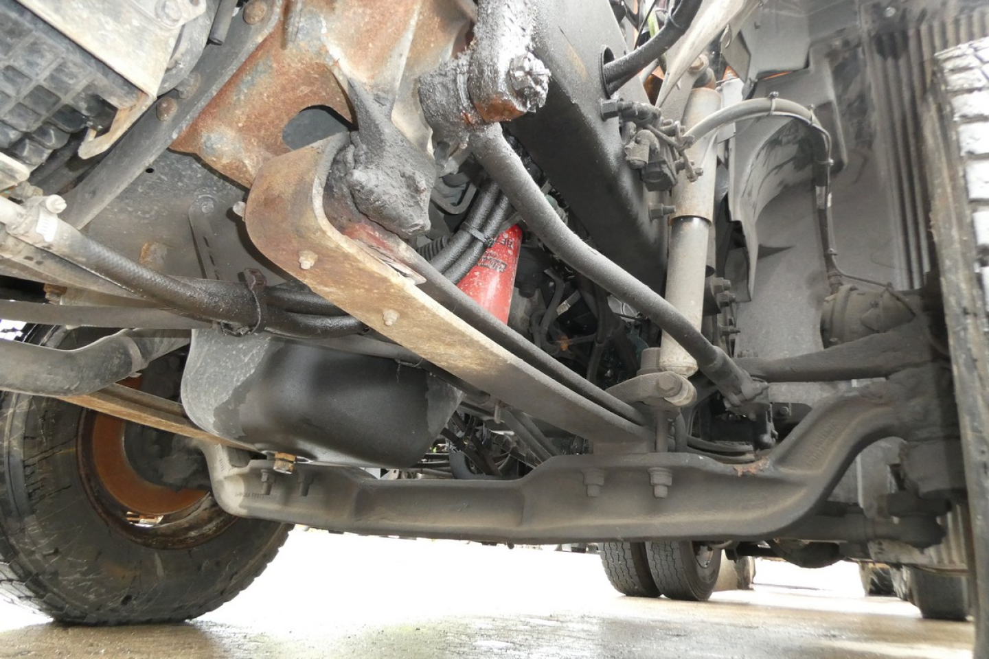 Truck undercarriage view showing suspension and steering components on a Volvo VNM42T. Visible are axle, leaf springs, tie rods, and frame with some rust and grime.