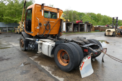 Orange Volvo VNM42T truck cab with single rear axle and exposed drivetrain, front view angled; visible wheels, fuel tank, and rear frame; background shows yard with equipment.