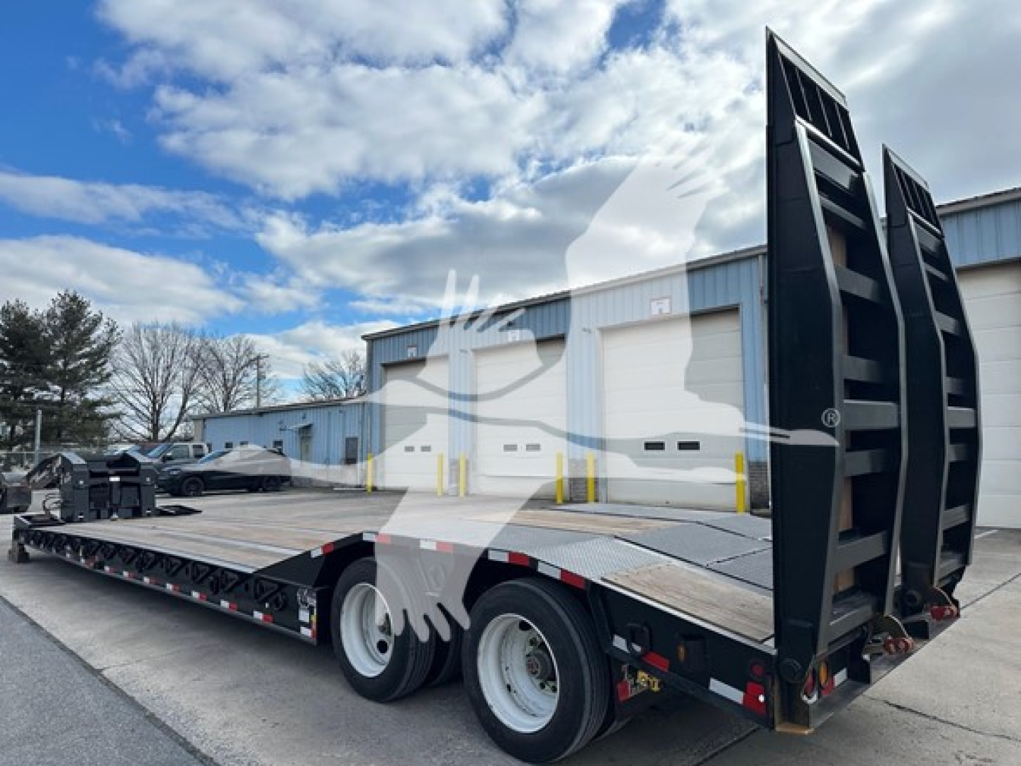 Lowboy trailer with extended flatbed and tall ramps, dual rear axles, and metal deck rails; side mirrors and blue sky visible.