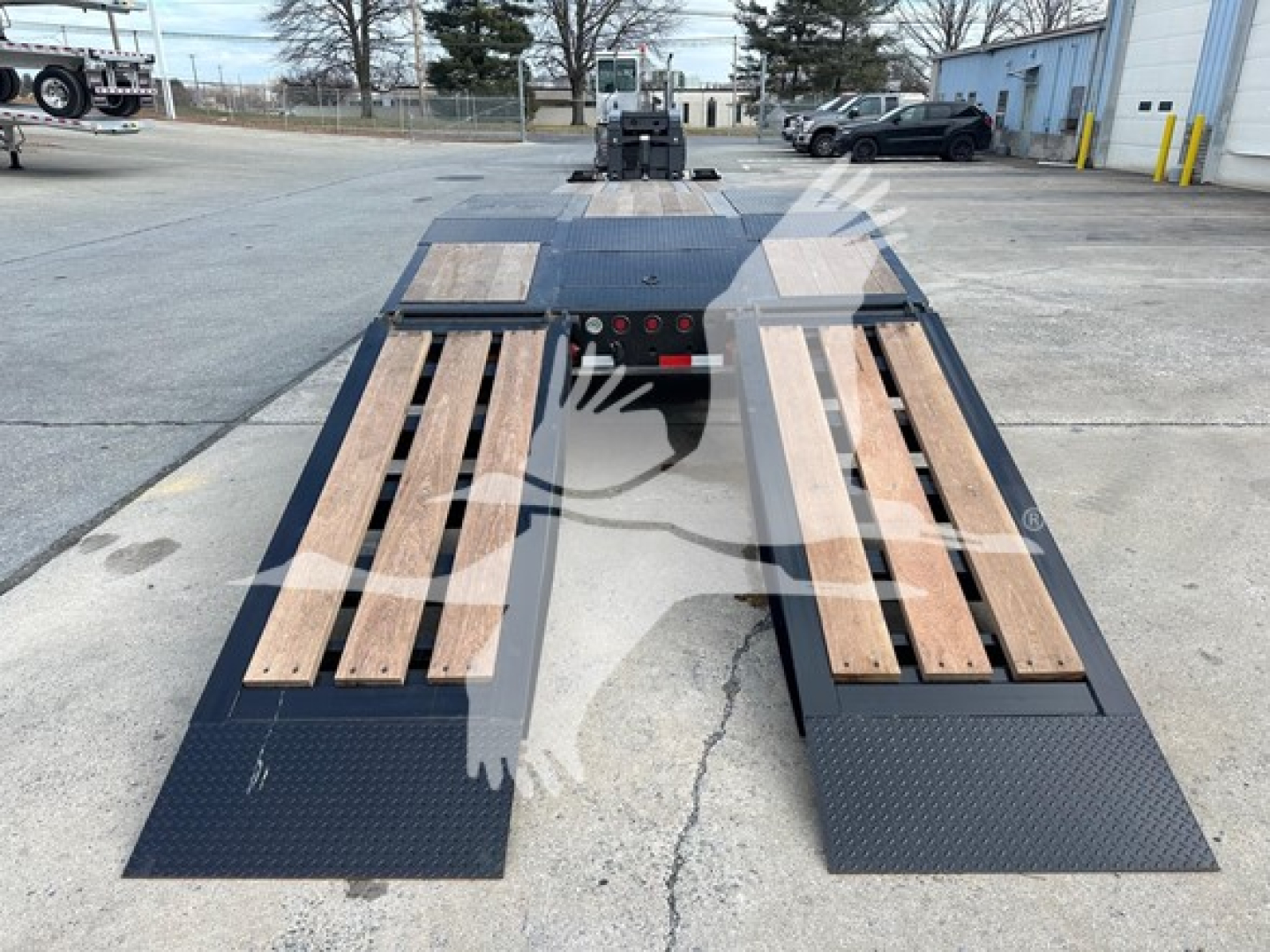 Lowboy trailer rear view showing dual loading ramps with wooden planks and diamond plate deck, blue/black metal surfaces.