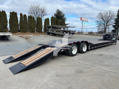 Lowboy trailer with folded drop-down ramps showing wooden deck and long dual-axle sections. Tail end view with black frame and wheelset, dealership lot in background.