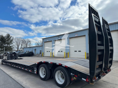 Trailer flatbed with two rear loading ramps up, 3-axle configuration, wooden deck, industrial building in background.