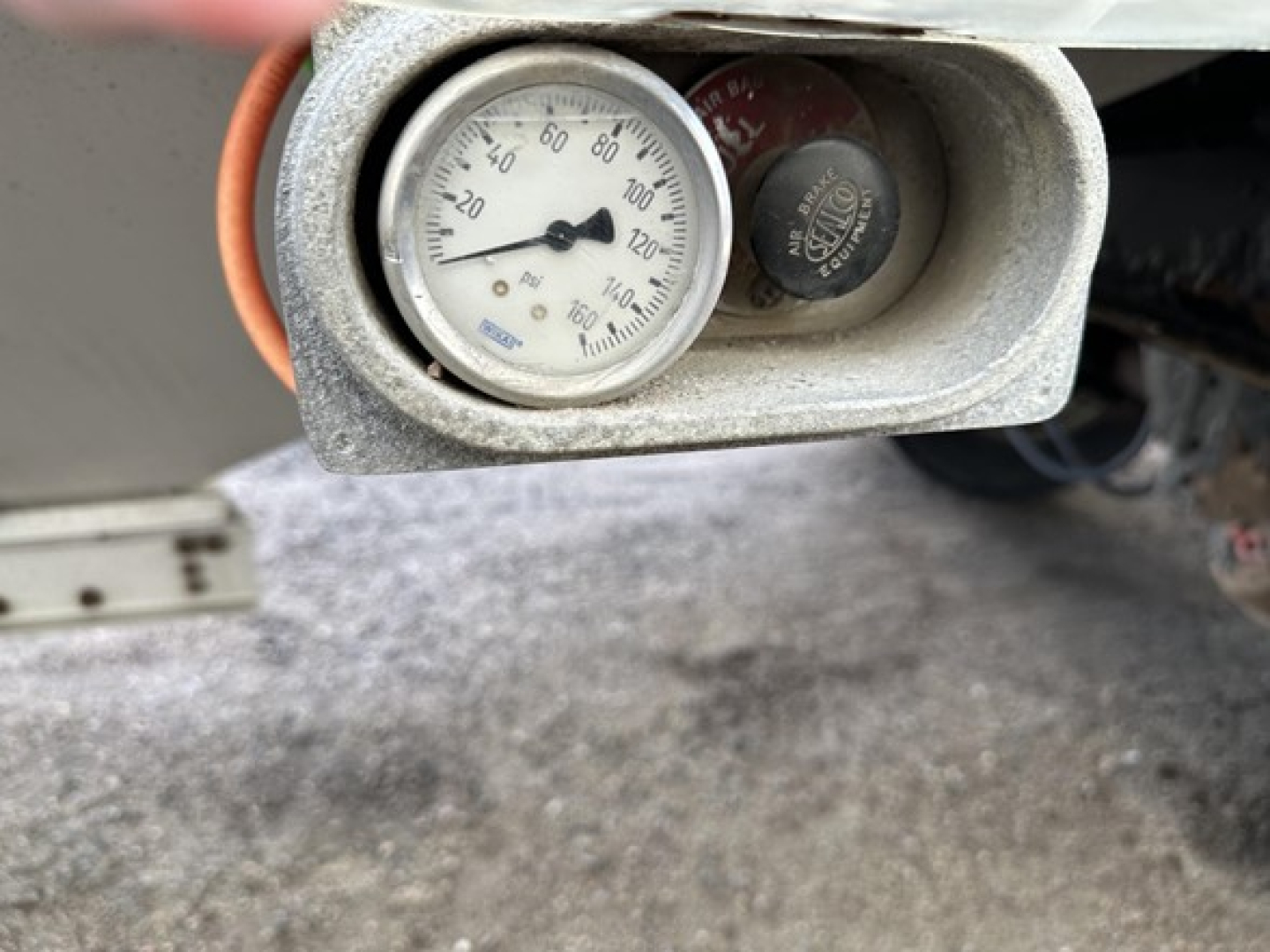 Trailer hitch gauge showing pressure dial and nearby valve cap; circular white gauge with numbers 0-140 and a pointer, mounted in gray housing.