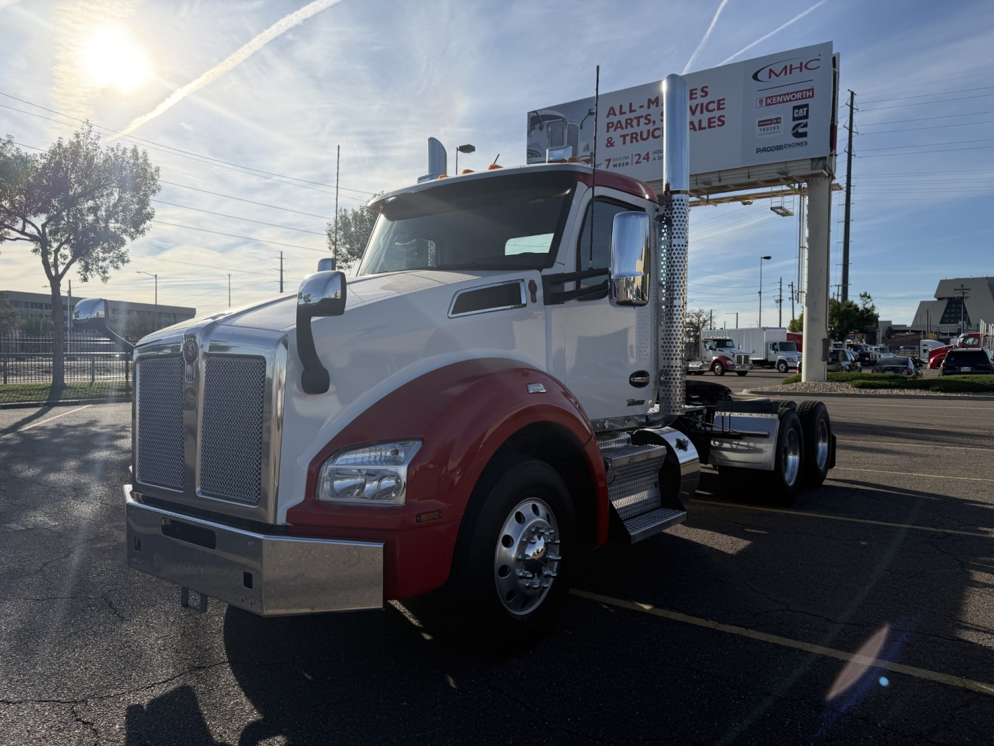 Kenworth T880 conventional daycab truck in a parking lot, front three-quarter view, white cab with red lower body, chrome grille, dual rear axles, 63k stock number sign in background.