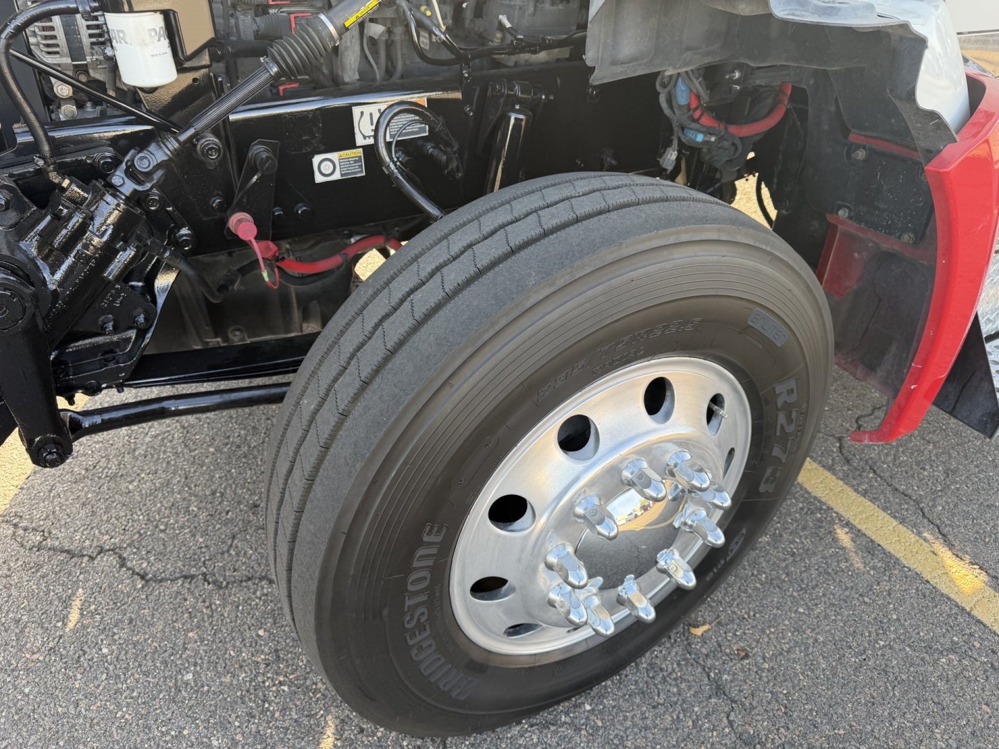 Kenworth T880 front wheel and suspension area showing Bridgestone tire and wheel hub; part of engine bay visible with hydraulic lines and components.