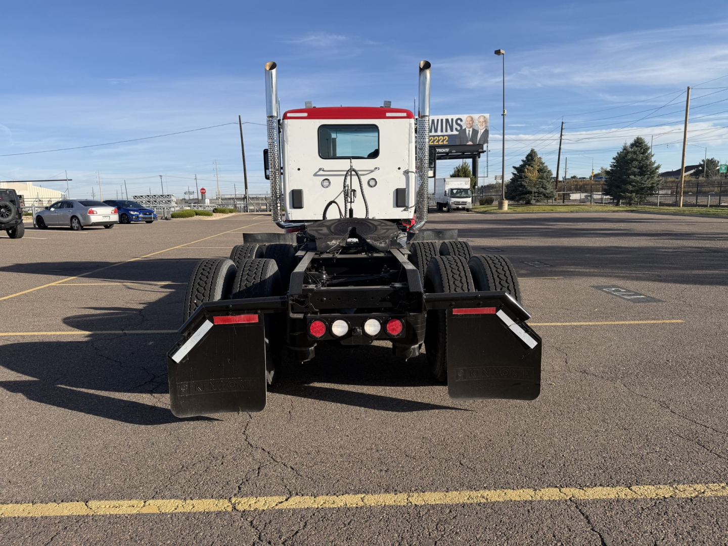 Kenworth T880 rear view showing two large mudflaps and dual rear wheels, fifth wheel and bumper area, white cab with red accents, in a parking lot.