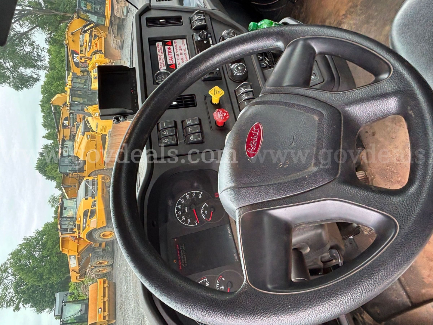 Peterbilt truck cockpit view showing steering wheel and dashboard with gauges; yellow heavy equipment vehicles visible through window.