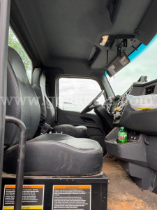Peterbilt truck cab interior showing steering wheel and dashboard with warning labels at footwell; driver and passenger seats visible; cab floor appears dusty.