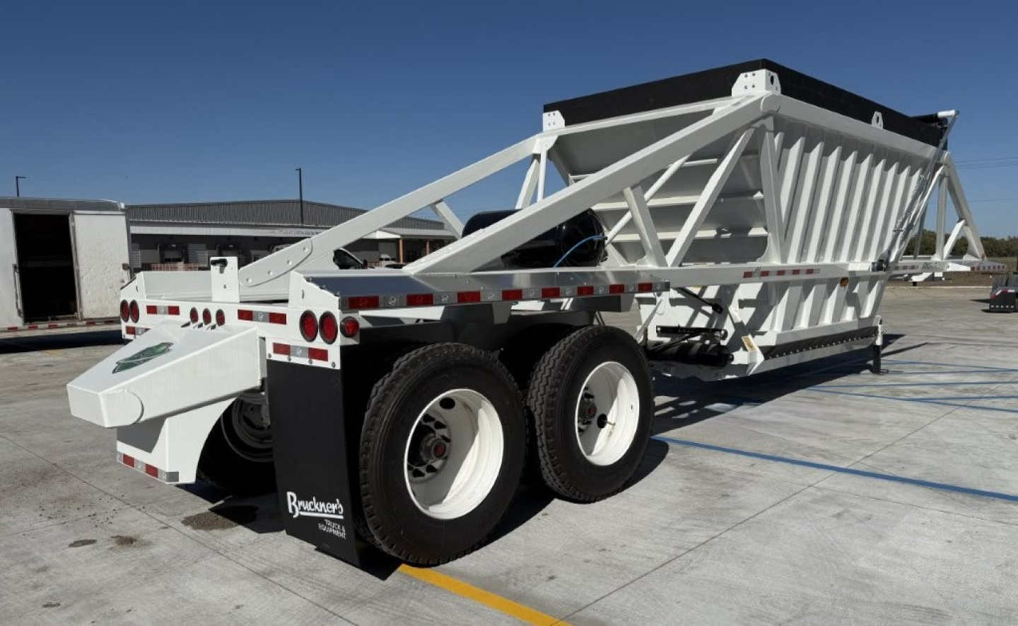 Dump trailer showing skeleton steel frame and dual rear axles with white wheels; long angled hitch and hopper on top.