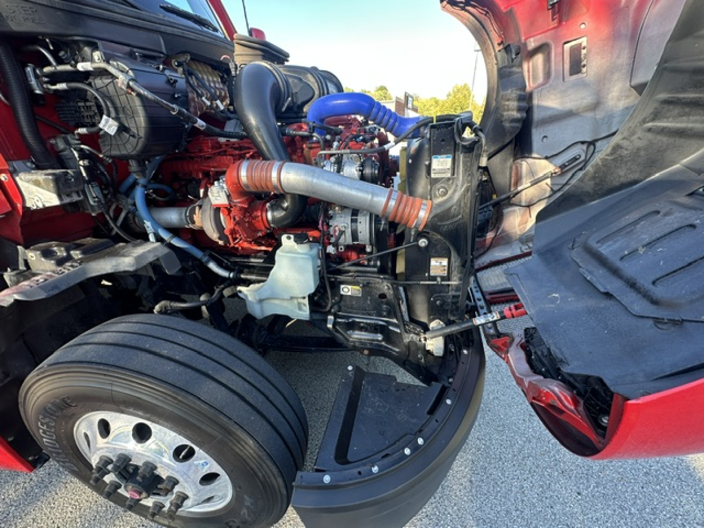 Kenworth T680 engine bay with red exterior showing air intake, hoses, and turbo; visible front wheel and open hood.