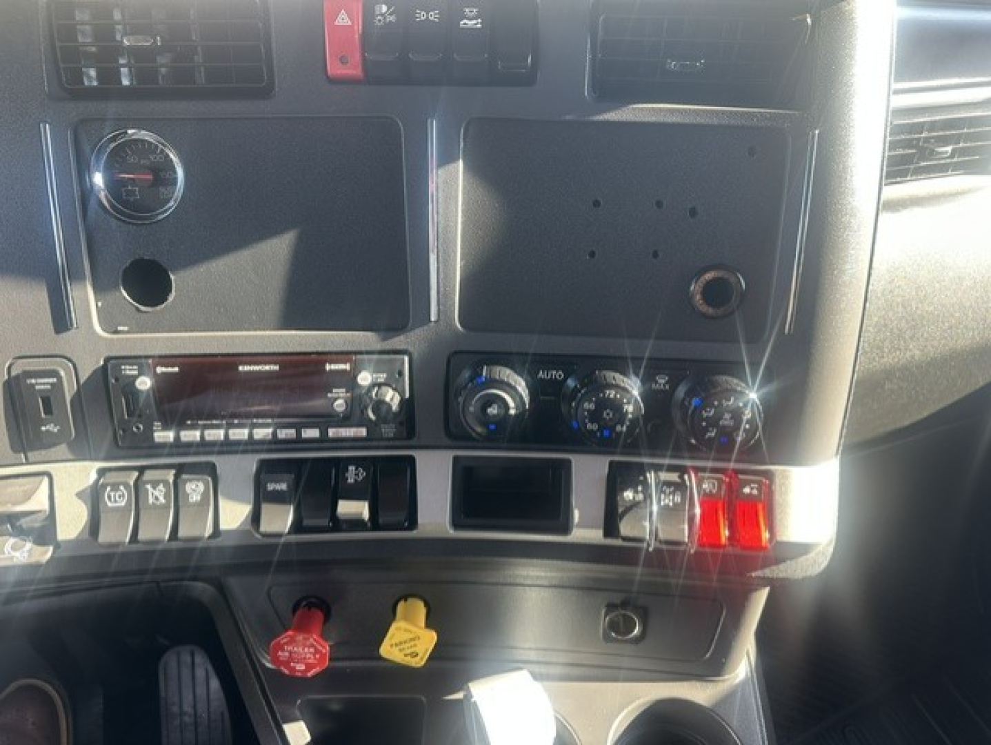 Kenworth truck interior dash showing climate controls, radio, and ignition area. Visible labels include red and yellow push buttons, blank panel, and various knobs.