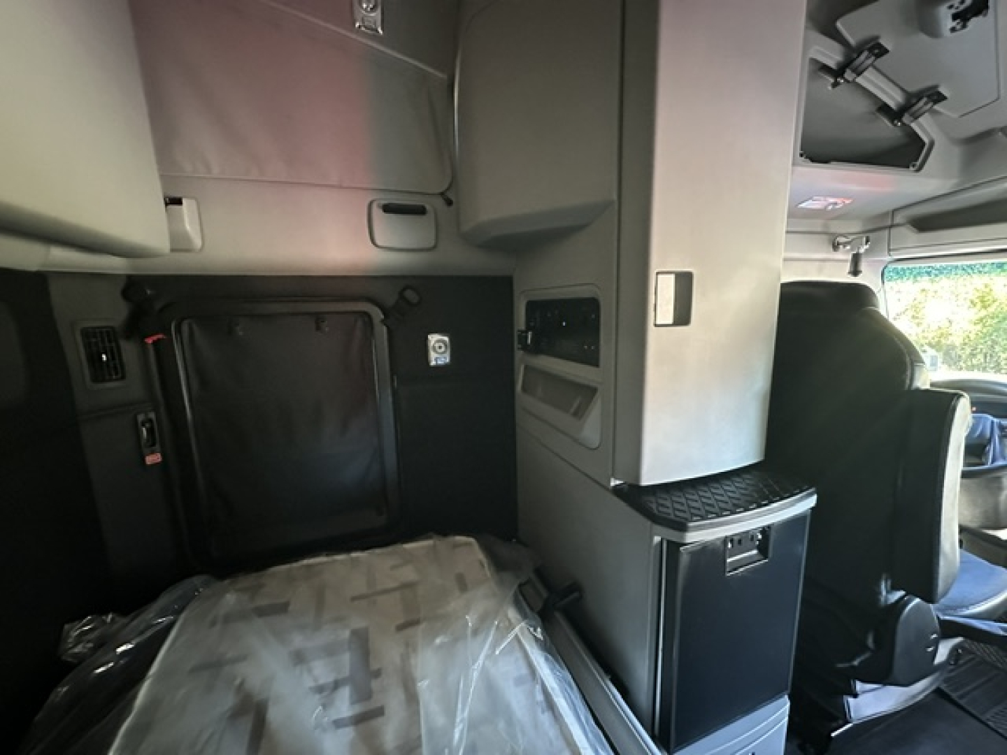 Truck interior showing sleeper area of a 2023 Kenworth T680 with overhead cabinets, black fabric mattress, and storage compartments in view
