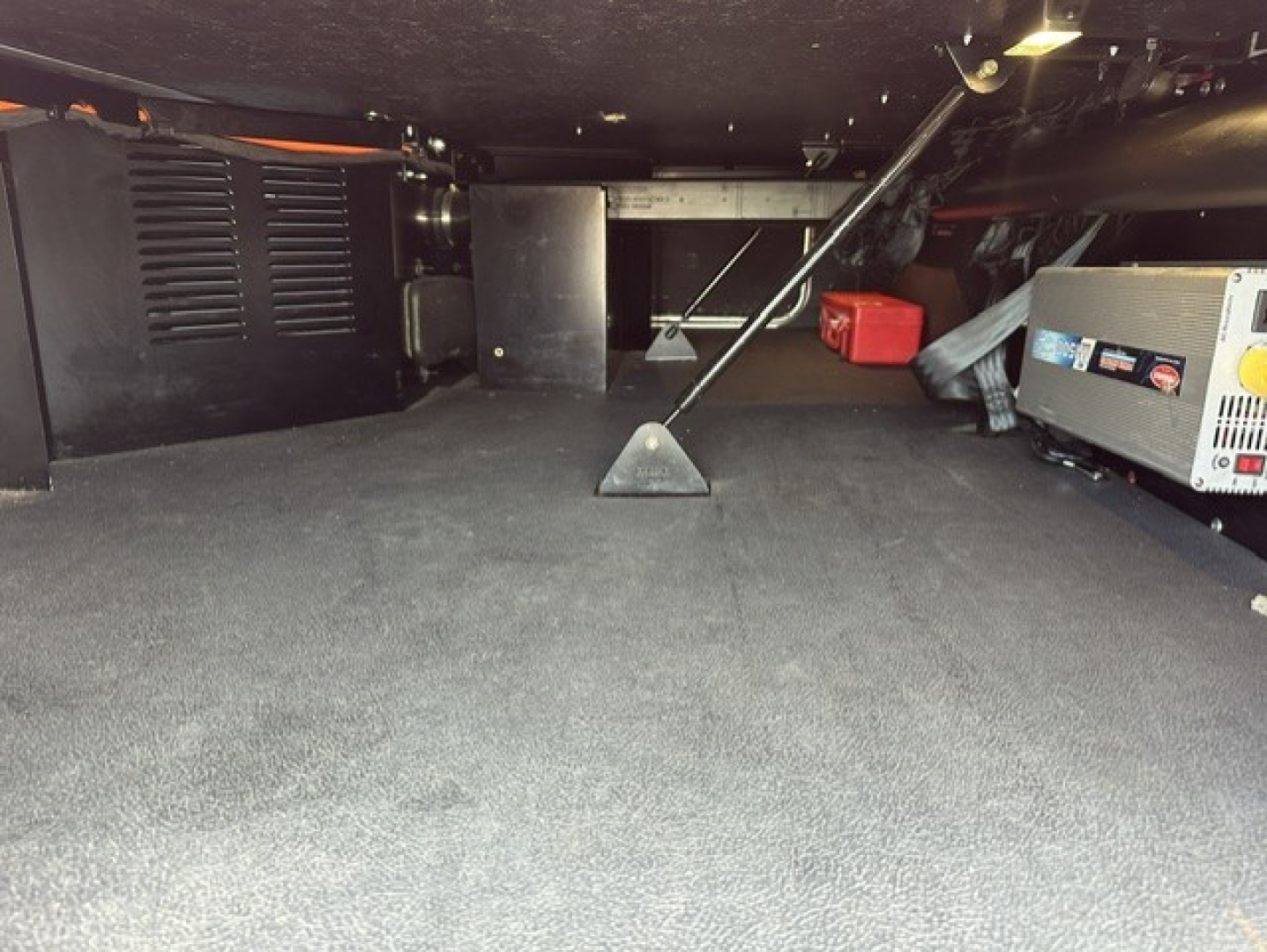 Interior undercarriage view of a 2023 Kenworth T680 sleeper showing a generator and storage area with a red tool box and black equipment, carpeted floor and support post in the middle.