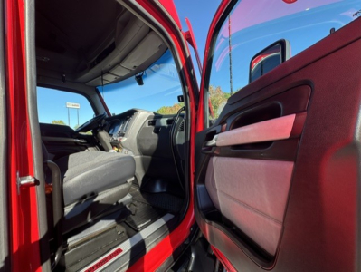 Front cabin of a red Kenworth T680 truck showing driver seat and open door with interior panels. Visible steering wheel and dashboard area.