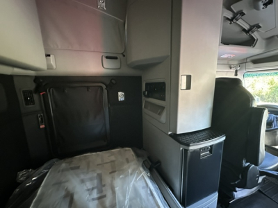 Truck interior showing sleeper area of a 2023 Kenworth T680 with overhead cabinets, black fabric mattress, and storage compartments in view