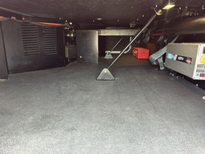 Interior undercarriage view of a 2023 Kenworth T680 sleeper showing a generator and storage area with a red tool box and black equipment, carpeted floor and support post in the middle.