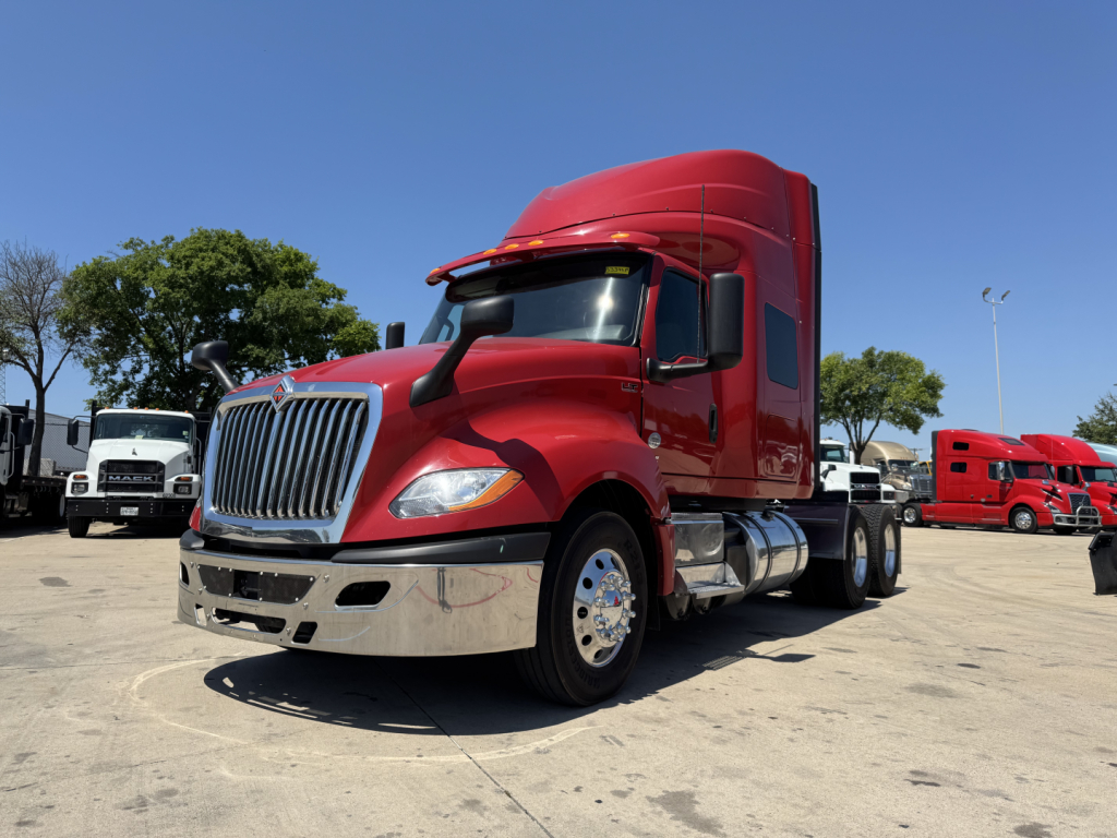 Truck cab in a dealership lot: 2022 International LT tractor with red cab, chrome front bumper, side mirrors, and sleeper cab visible; other trucks in background.