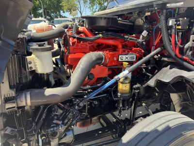 Truck engine bay showing a bright red Cummins X15 engine with surrounding hoses, reservoirs, and components in view.