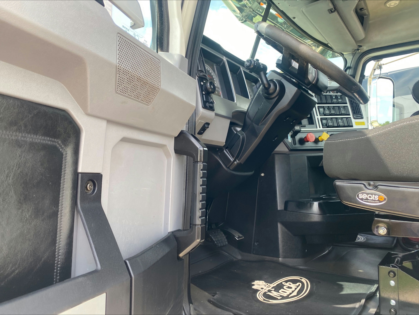 Interior front cab of a Mack PI64T truck showing steering wheel, dashboard, and driver seat with Seots branding; floor mat shows Mack logo.