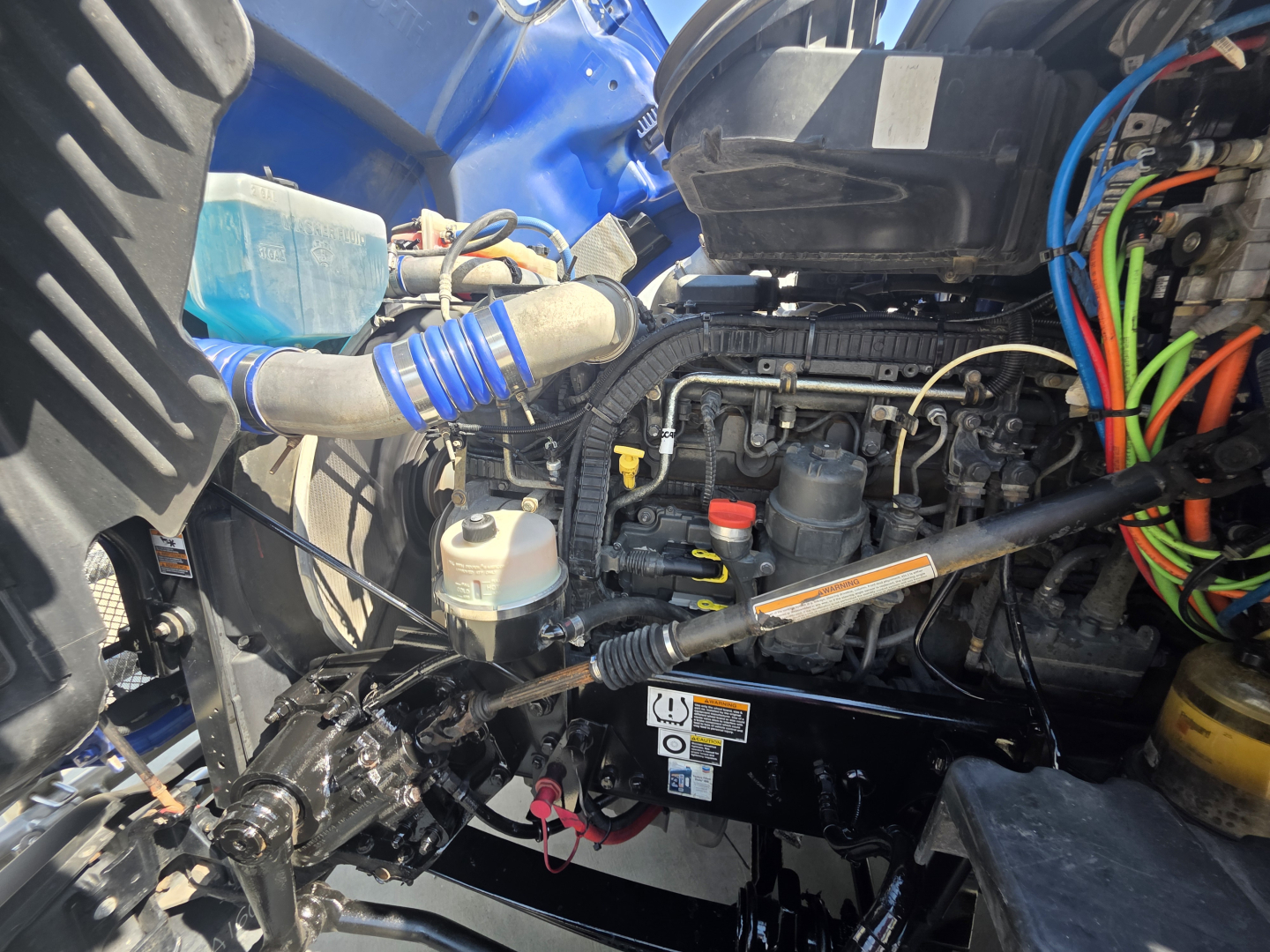 Kenworth T680 engine bay with various hoses and wiring; visible components include coolant reservoir, blue coolant hose, electrical harnesses, and engine block.
