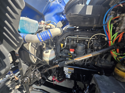 Kenworth T680 engine bay with various hoses and wiring; visible components include coolant reservoir, blue coolant hose, electrical harnesses, and engine block.