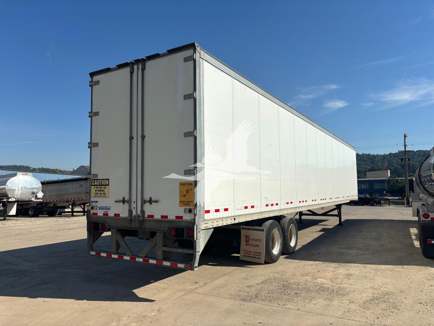 Trailer rear and side view showing white enclosed Vanguard VXP trailer with dual-axle configuration and rear doors, CAUTION labels on doors, reflective tape along lower edge.