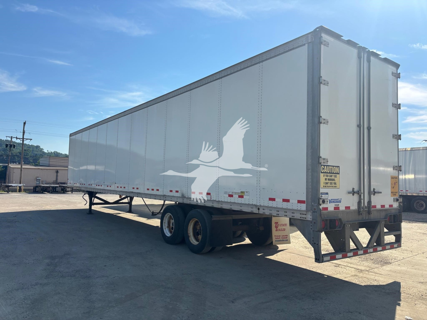 Trailer side view of a Vanguard VXP box trailer with a large white graphic of a flying crane. Dual rear doors, side marker lights, and labeling/CAUTION sticker near the back.