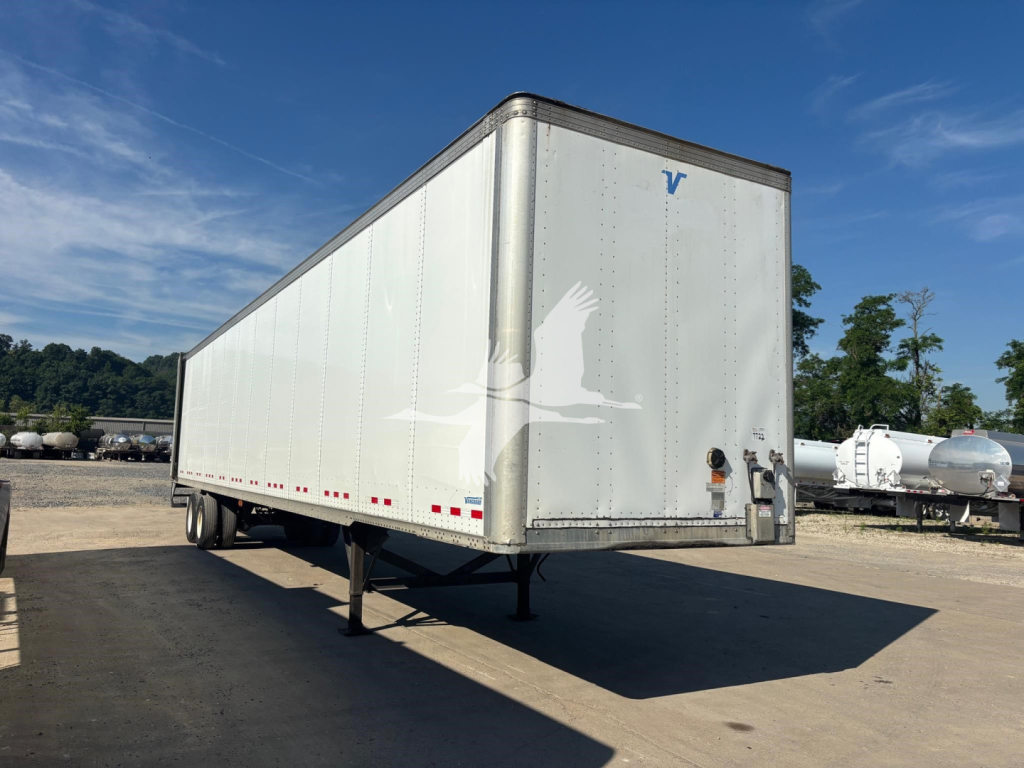 Vanguard VXP trailer in a yard showing a plain silver exterior with a small blue Vanguard logo on the front corner and wheels tucked under, side view.