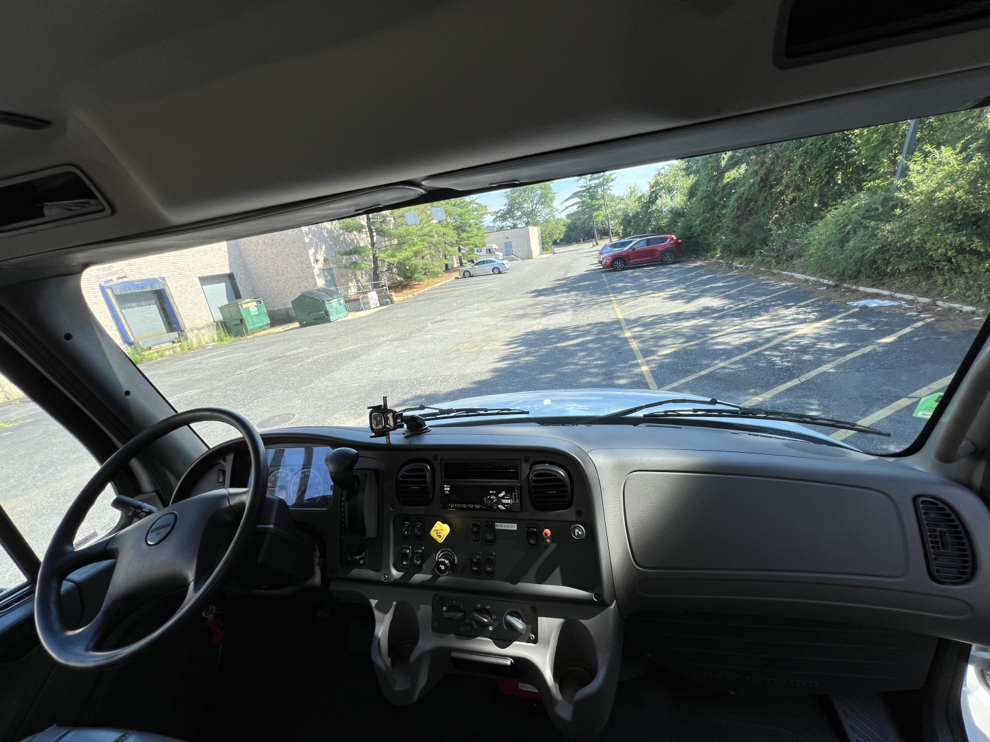 Cab interior of a 2019 Freightliner M2 Series truck showing dashboard, steering wheel, and driver’s seat with view through windshield to a driveway.