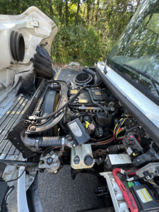 Freightliner M2 series engine bay showing front of cab with exposed engine components, wiring, hoses, and battery in view.