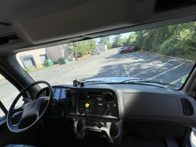 Cab interior of a 2019 Freightliner M2 Series truck showing dashboard, steering wheel, and driver’s seat with view through windshield to a driveway.