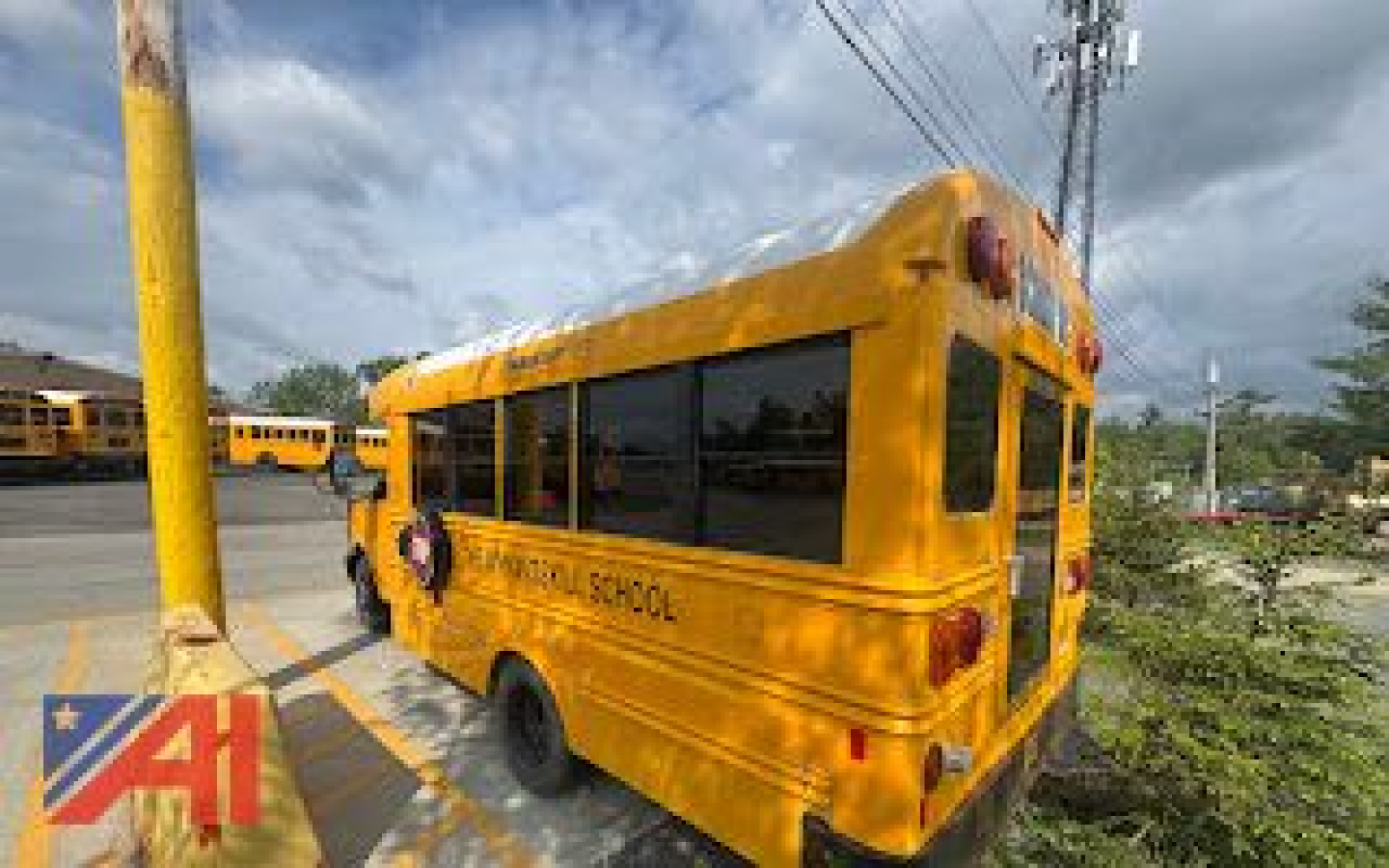 Rear/side view of a yellow school bus with black windows, text 'SCHOOL' along the side and rear, taillights visible, utility pole and other yellow buses in background.