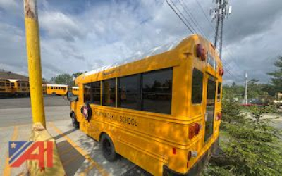 Rear/side view of a yellow school bus with black windows, text 'SCHOOL' along the side and rear, taillights visible, utility pole and other yellow buses in background.